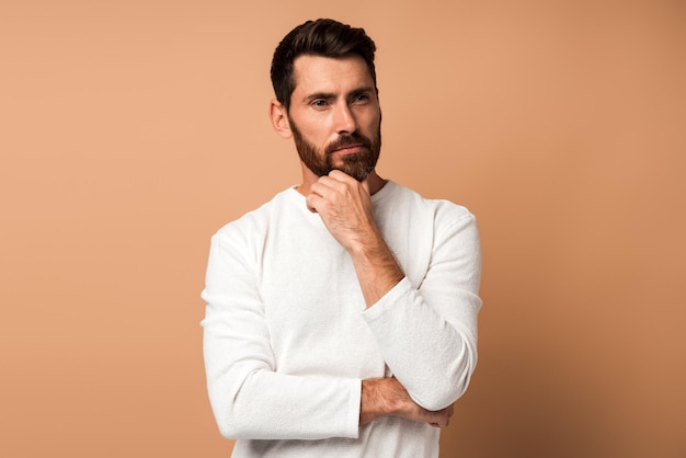 Necesito pensar Retrato de un hombre moreno pensativo con barba en camiseta blanca sosteniendo su barbilla y reflexionando sobre una idea confundida, no estoy seguro acerca de la solución, una toma de estudio interior aislada en un fondo beige