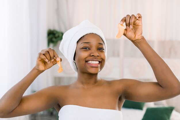 Necesito un poco de colágeno. Atractiva joven africana en una toalla blanca aplicando parche en el ojo médico y sonriendo mientras está de pie contra el fondo gris