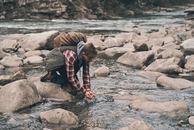 Necesito refresco. Apuesto joven moderno bebiendo agua del río mientras camina en las montañas