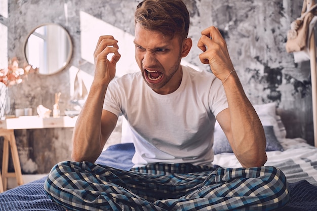 Negativ begeisterter brünette Mann, der Fäuste macht, während er seine Gefühle demonstriert und auf dem Bett sitzt. Hauskonzept