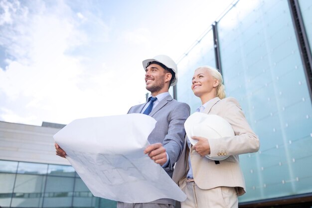 negocio, asociación, arquitectura y concepto de personas - hombre de negocios sonriente y mujer de negocios con planos y cascos en la calle de la ciudad