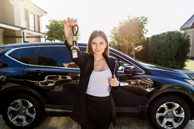 Foto negocio automotriz, venta de automóviles, gesto y concepto de personas: feliz empresaria o vendedora caucásica con carpeta dando la clave del automóvil sobre el fondo del automóvil nuevo al aire libre en el salón del automóvil