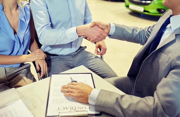 Foto negocio de automóviles, venta de automóviles, gestos y concepto de personas: cierre de la pareja de clientes con el distribuidor dándose la mano en el salón de automóviles o en el salón