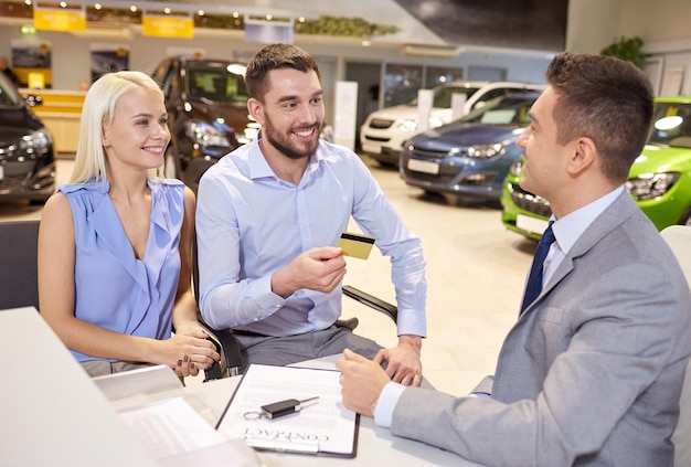 negocio de automóviles, venta y concepto de personas - pareja feliz con un concesionario que compra un automóvil en una exhibición de automóviles o en un salón