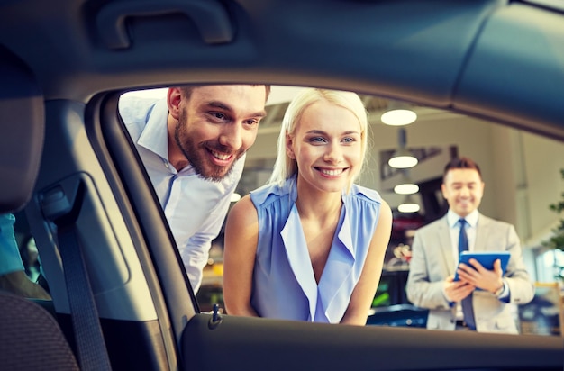 negocio de autos, venta de autos, tecnología y concepto de personas - pareja feliz con concesionario de autos en exhibición de autos o salón