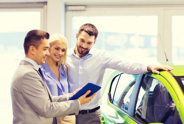 negocio de autos, venta de autos, tecnología y concepto de personas - pareja feliz con concesionario de autos en exhibición de autos o salón