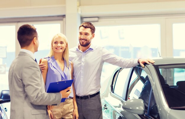 negocio de autos, venta de autos, tecnología y concepto de personas - pareja feliz con concesionario de autos en exhibición de autos o salón