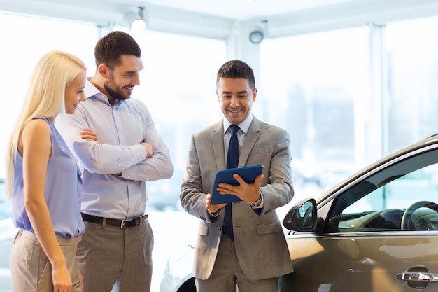 negocio de autos, venta de autos, tecnología y concepto de personas - pareja feliz con concesionario de autos en exhibición de autos o salón