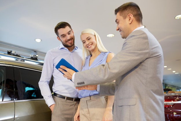 negocio de autos, venta de autos, tecnología y concepto de personas - pareja feliz con concesionario de autos en exhibición de autos o salón