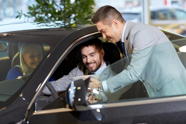 negocio de autos, venta de autos, tecnología y concepto de personas - pareja feliz con concesionario de autos en exhibición de autos o salón