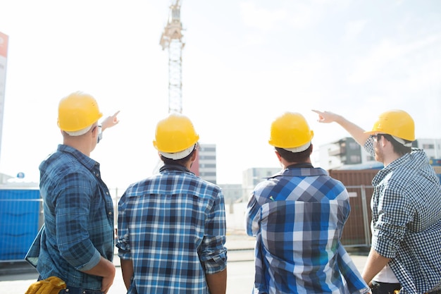 negocio, construcción, trabajo en equipo, desarrollo y concepto de personas - grupo de constructores en cascos en el sitio de construcción apuntando con el dedo a la grúa desde atrás