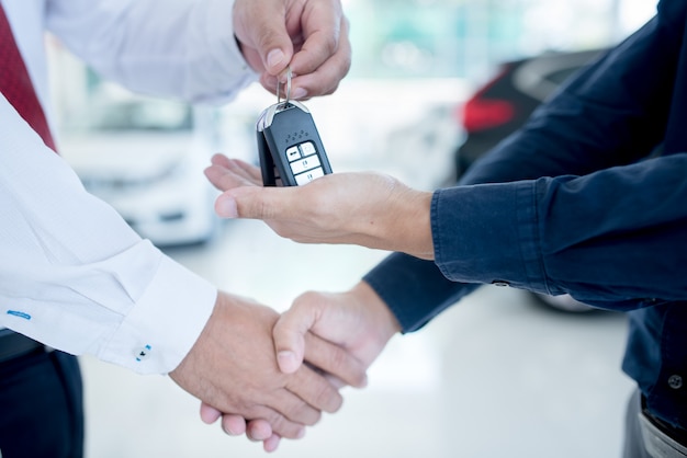 negócio de automóveis, venda de carros, acordo, gesto e conceito de pessoas - close-up de revendedor, dando a chave ao novo proprietário e apertando as mãos no salão do automóvel ou salão de beleza