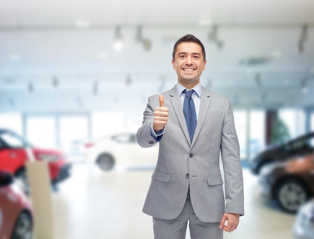 negócio de automóveis, venda de carros, consumismo, gesto e conceito de pessoas - homem feliz mostrando os polegares sobre o salão de automóveis ou fundo de salão