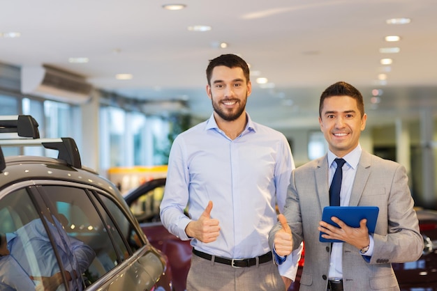 negócio de automóveis, venda de carros, tecnologia, gesto e conceito de pessoas - homem feliz e revendedor de automóveis com computador tablet pc mostrando os polegares no salão de automóveis ou salão