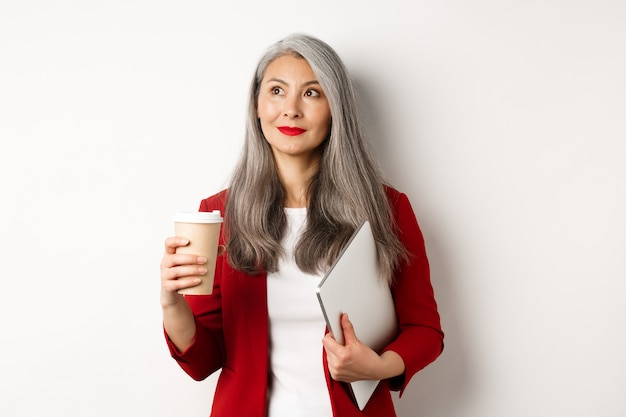 Negocio. Exitosa empresaria asiática con cabello gris, tomando café y de pie con el portátil, mirando pensativo en la esquina superior izquierda.