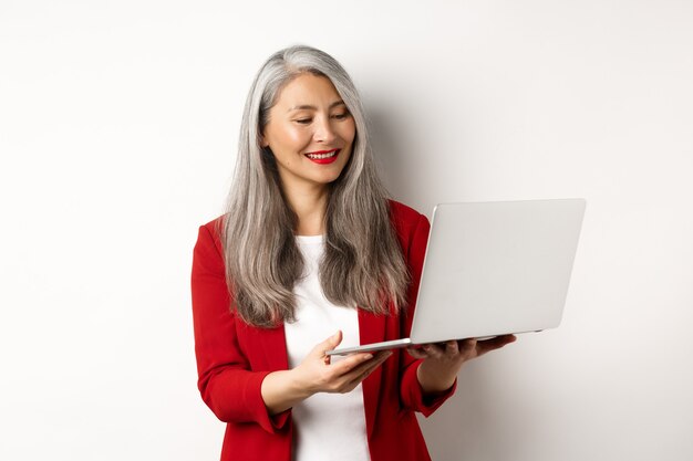 Negocio. Exitosa empresaria asiática en chaqueta roja trabajando en la computadora portátil, sonriendo y leyendo la pantalla, de pie sobre fondo blanco.