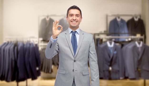 negocio, gente, ropa de hombre, venta y concepto de ropa - hombre de negocios sonriente feliz en traje sobre el fondo de la tienda de ropa que muestra el signo de la mano ok