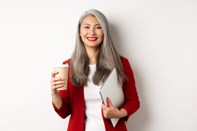 Negocio. Gerente femenina asiática exitosa de pie con una taza de café y un portátil, sonriendo satisfecho ante la cámara, fondo blanco.