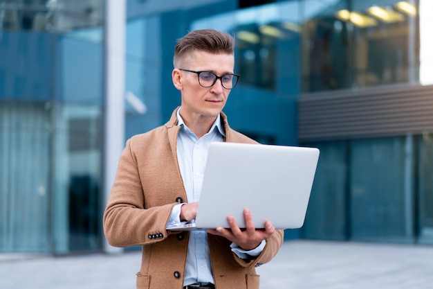 Negocio. Hombre de negocios que usa la computadora portátil al aire libre Adulto Caucásico Masculino Persona de negocios Anteojos Mirando la pantalla del portátil Afuera Pensamiento serio Fondo de luz solar Calle de la gran ciudad