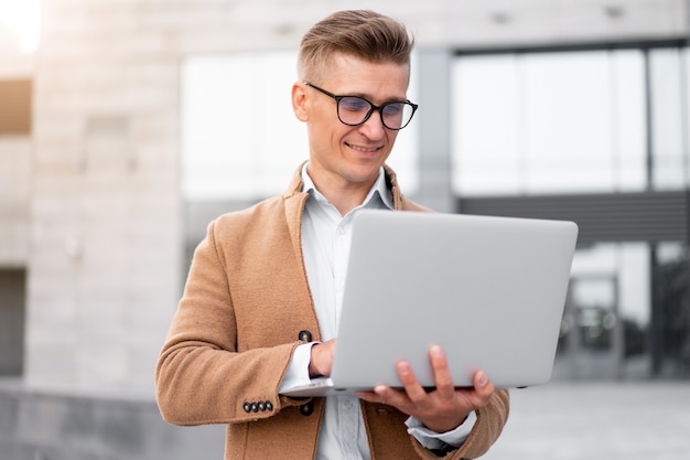 Negocio. Hombre de negocios que usa la computadora portátil al aire libre Adulto Caucásico Masculino Persona de negocios Anteojos Vestida Chaqueta Hipster Afuera Sonriendo Feliz Emociones positivas Luz del sol Fondo Calle de la gran ciudad