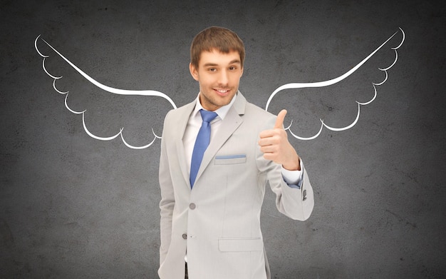 Foto negocio, inversionista ángel, seguridad, seguridad y concepto de personas - joven hombre de negocios sonriente con alas dibujando mostrando los pulgares hacia arriba sobre fondo de hormigón gris