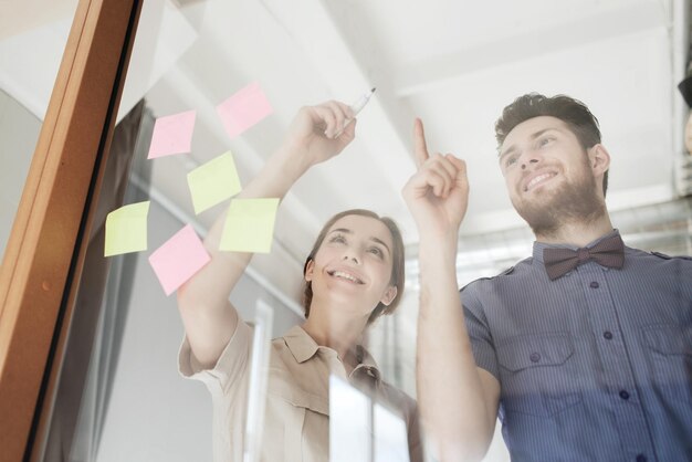 negocio, puesta en marcha, planificación, gestión y concepto de personas - equipo creativo feliz escribiendo o dibujando algo en una pizarra de vidrio de oficina en blanco