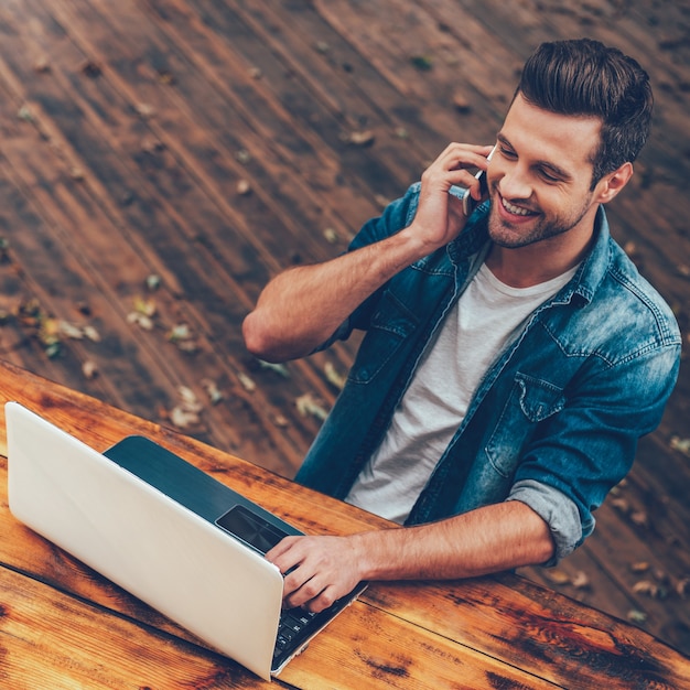 Negócios ao ar livre. Vista superior de um jovem feliz trabalhando em um laptop e falando ao telefone móvel enquanto está sentado à mesa de madeira ao ar livre
