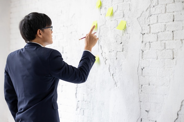 Negocios asiáticos en anteojos se encuentra en la ventana, use notas adhesivas, horario de recordatorio de papel para discutir la idea en la oficina, empresario mirando y pensando en el cuaderno de notas en la pared blanca.