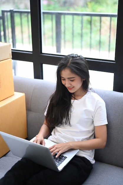 Negocios asiáticos jóvenes ponen en marcha el propietario del vendedor en línea usando una computadora portátil para verificar los pedidos de los clientes desde el sitio web mientras están sentados en el sofá.