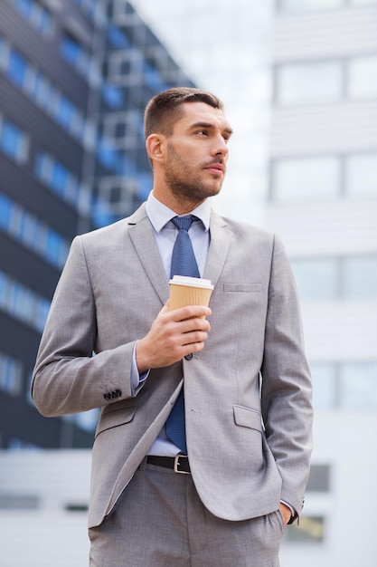 negócios, bebidas quentes e pessoas e conceito - jovem empresário sério com xícara de café de papel sobre prédio de escritórios