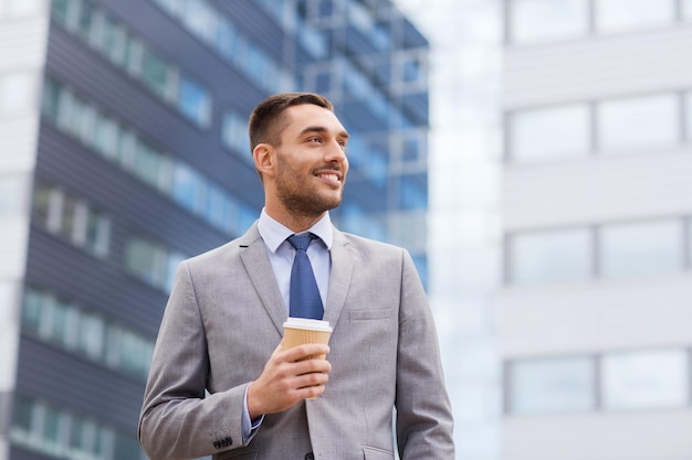 negócios, bebidas quentes e pessoas e conceito - jovem empresário sorridente com xícara de café de papel sobre prédio de escritórios