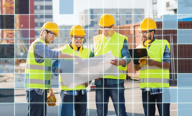 negocios, construcción, trabajo en equipo, tecnología y concepto de personas - grupo de constructores sonrientes con cascos y chalecos muy visibles mirando planos y portapapeles al aire libre sobre fondo de cuadrícula cuadrada