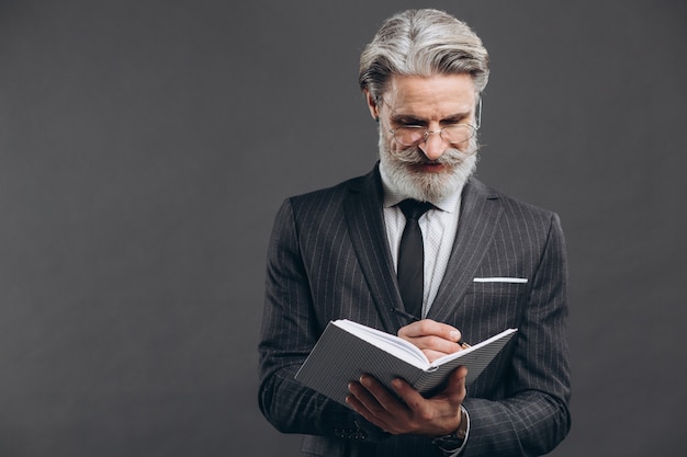 Negócios e elegante homem maduro barbudo em um terno cinza, escrevendo no seu espaço de cópia de caderno na parede cinza.