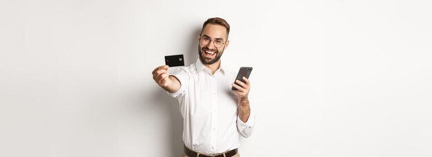 Negócios e pagamento online animado homem mostrando seu cartão de crédito enquanto segura o smartphone em pé sa