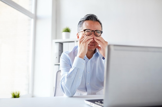 Negocios, exceso de trabajo, fecha límite y concepto de personas: hombre de negocios cansado con anteojos y computadora portátil frotándose los ojos en la oficina