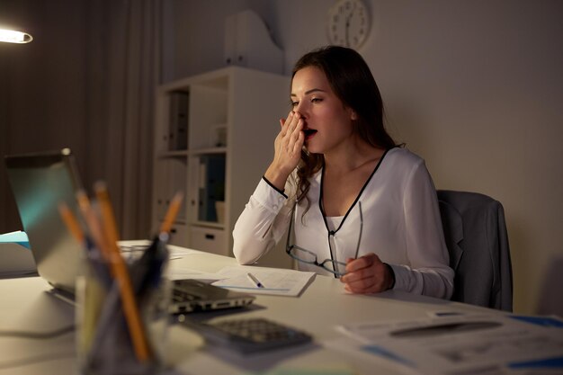 negocios, exceso de trabajo, plazo y concepto de personas - mujer cansada con papeles y computadora portátil bostezando en la oficina nocturna