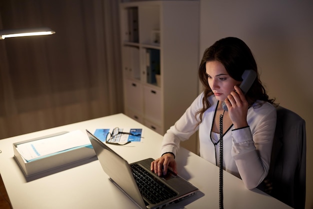 negocios, exceso de trabajo, plazo y concepto de personas - mujer escribiendo en la computadora portátil y llamando por teléfono en la oficina nocturna