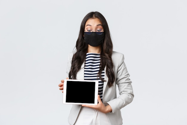 Negocios, finanzas y empleo, covid-19 previniendo el virus y el concepto de distanciamiento social. Trabajador de oficina asiático impresionado en mascarilla y gafas mirando sorprendido, mostrando la pantalla de la tableta digital