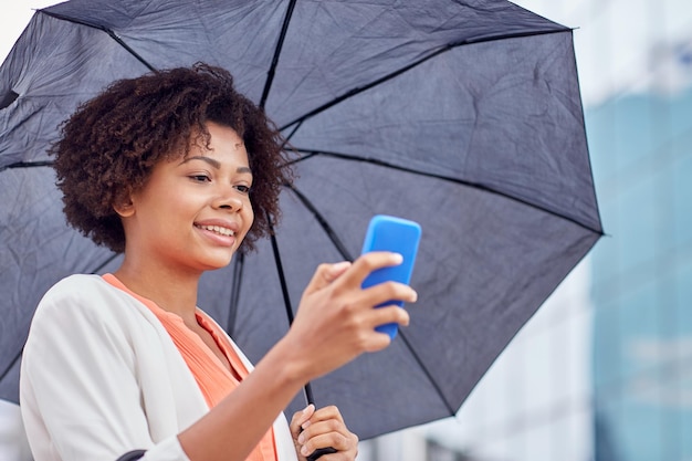 negocios, mal tiempo y gente y concepto - feliz mujer de negocios afroamericana con paraguas enviando mensajes de texto en un smartphone en la calle de la ciudad