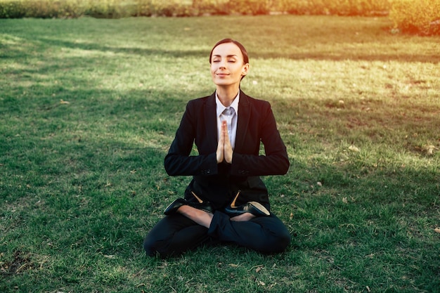 Negocios y meditación Una hermosa mujer de negocios concentrada con traje negro está sentada en una posición de loto en el césped del parque de la ciudad y descansando después del trabajo