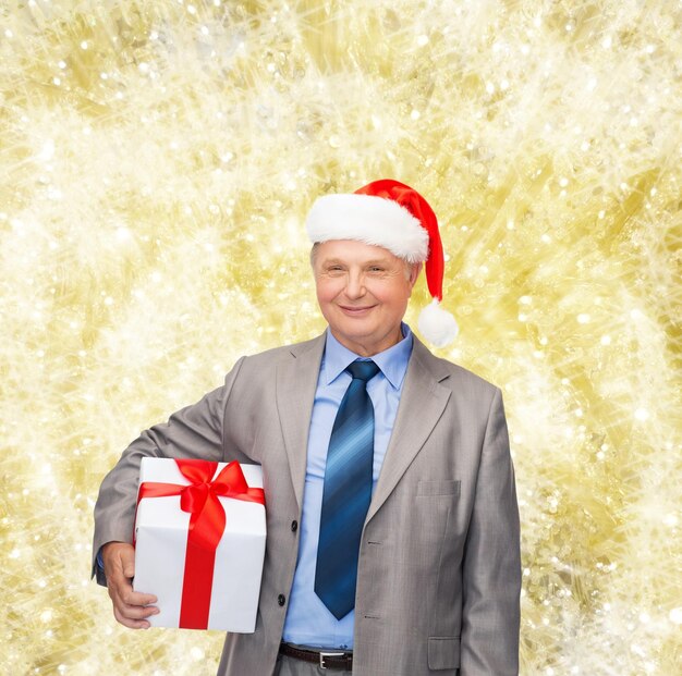 negocios, navidad, regalos y concepto de personas - anciano sonriente con traje y sombrero de ayudante de santa con regalo sobre fondo de luces amarillas
