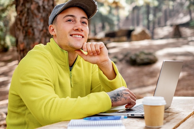 Negócios no ar fresco Jovem sorridente bonito trabalhando no laptop enquanto está sentado na mesa de madeira ao ar livre na floresta Conceito de trabalho inteligente na natureza