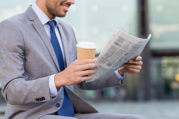 negocios, noticias, descanso y gente y concepto - cerca de un hombre de negocios sonriente leyendo el periódico y bebiendo café de un vaso de papel sobre el edificio de oficinas