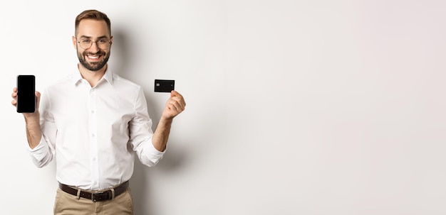 Foto negocios y pago en línea hombre guapo sonriente que muestra la pantalla del móvil y la tarjeta de crédito de pie