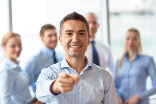 negocios, personas, gestos y concepto de trabajo en equipo: un hombre de negocios sonriente señalándote con un grupo de empresarios reunidos en el cargo