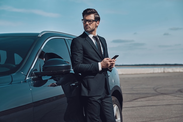 Negócios são sua vida. Belo jovem empresário usando um telefone inteligente enquanto está perto de seu carro ao ar livre