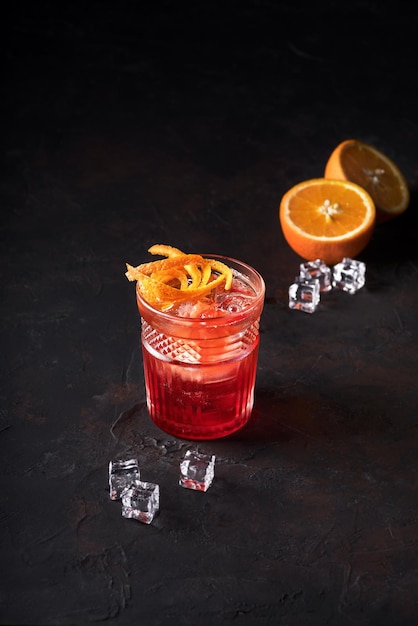 Negroni cocktail com orange twist e pin em fundo escuro e ao lado laranja e gelo fotografia de bebidas