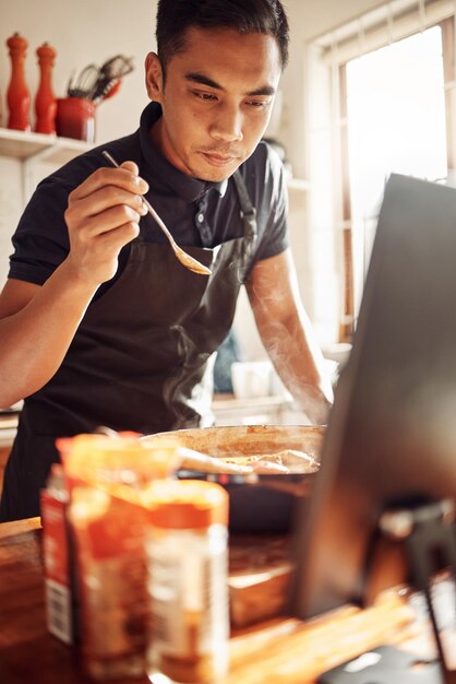 Nehmen Sie bequem von zu Hause aus an einem Kochkurs teil Aufnahme eines jungen Mannes, der einen Laptop verwendet, während er zu Hause eine Mahlzeit zubereitet