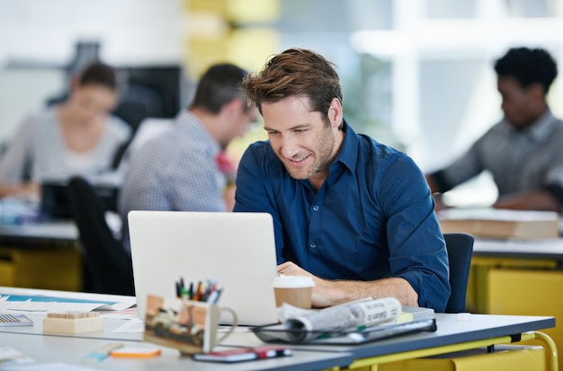 Nehmen Sie jedes Projekt in Angriff. Aufnahme eines Designers, der an seinem Schreibtisch sitzt und mit Kollegen im Hintergrund an einem Laptop arbeitet