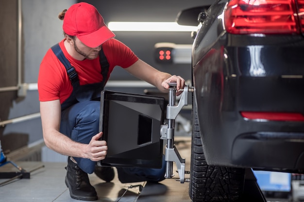 Neigungswinkelausrichtung. Junger erwachsener Mann in der Arbeitskleidung, die Rad begradigt, hockte nahe Auto in Werkstatt